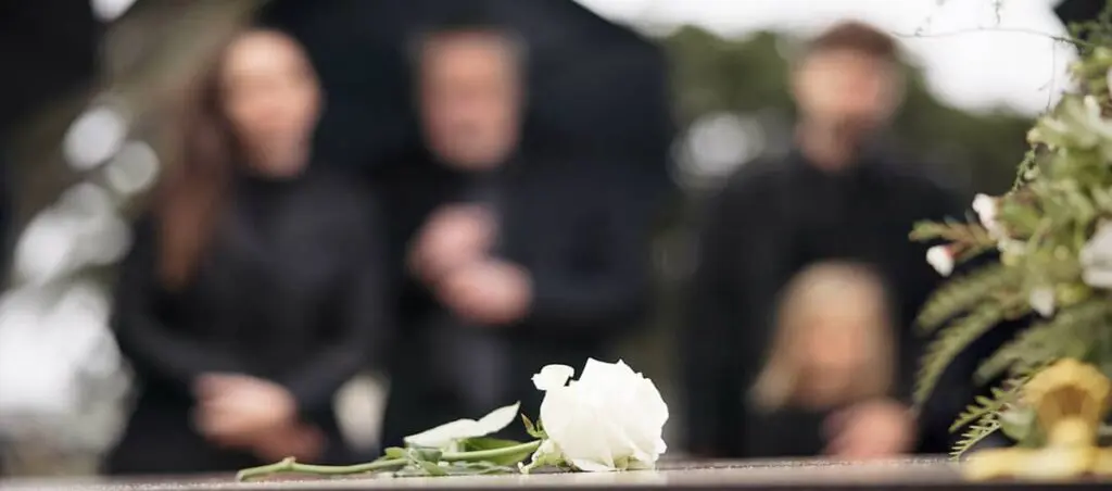 A white rose rests on a coffin with a blurred audience in the background. This illustrates the concept of performing a funeral for strangers.