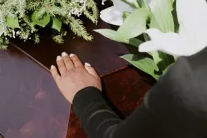 A hand rests on a coffin; some greenery shows at the top of the photo.