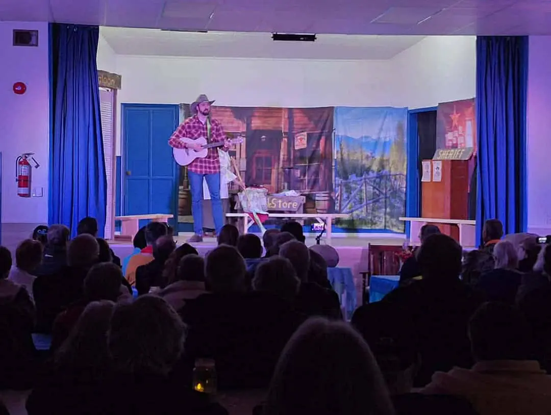 Jay Foster holds a guitar while standing on a stage in front of an audience during a community drama group performance.