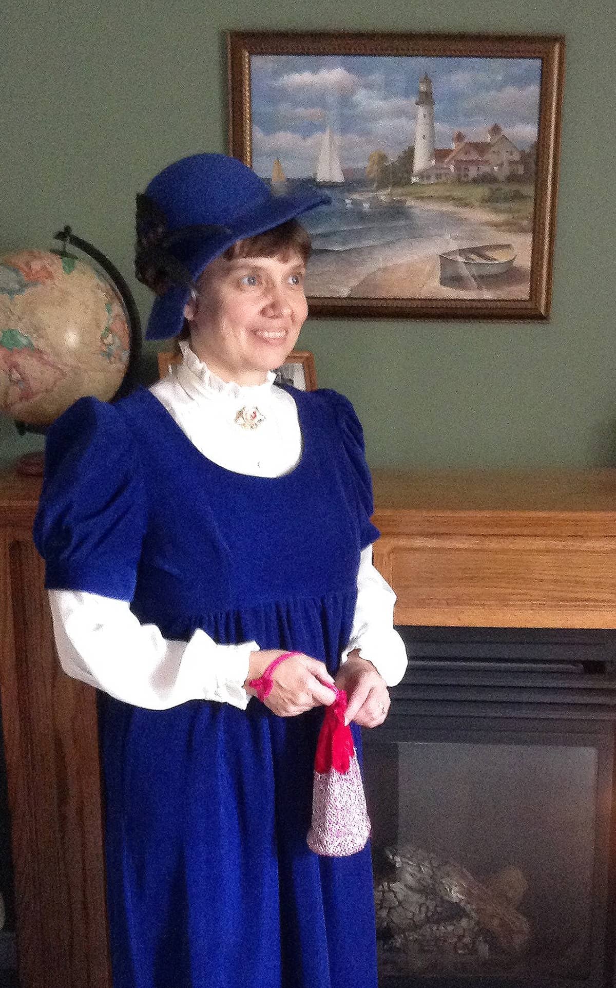 Joy Deneault wears an old-fashioned blue dress, along with matching hat and carries a red drawstring bag.