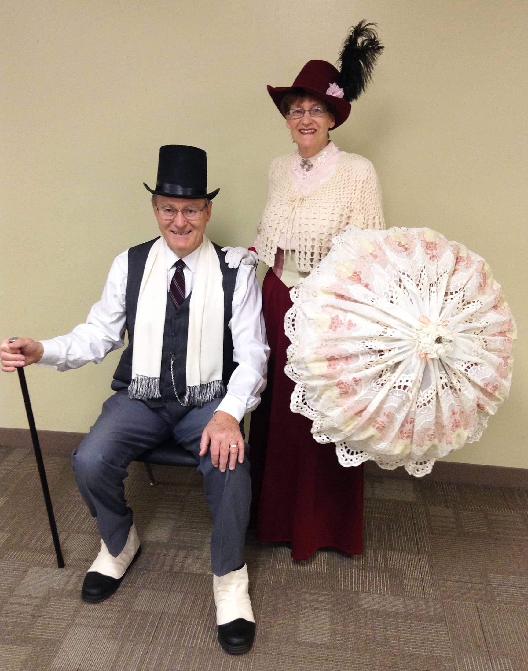 Bill & Lorna Powell wear old-fashioned clothing; Bill has a walking cane and top hat, while Lorna carries a parasol and wears a hat with a feather in it.